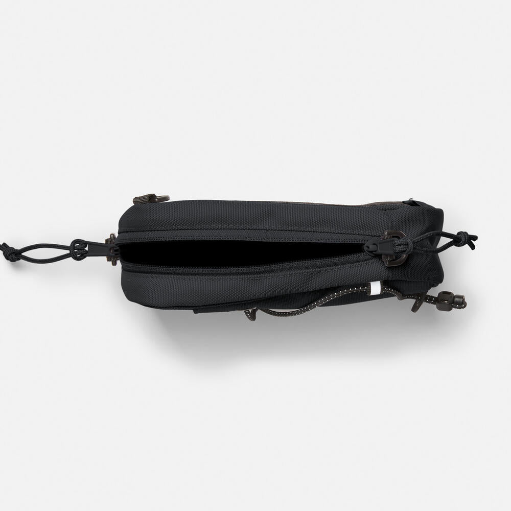 Timberland® Outdoor Archive 2.0 Crossbody Bag in Black. Black crossbody bag made from 100% recycled polyester. Features a large zipper opening, front panel slip pocket with bungee system, adjustable shoulder strap, and two ways to wear. Compact size and lightweight design. Sustainable choice for everyday use.